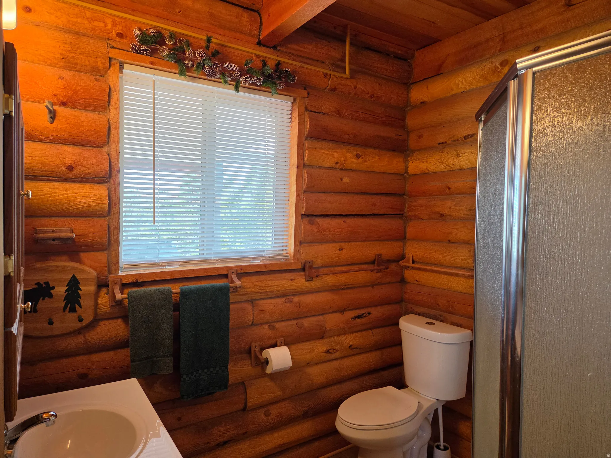 Wood-paneled walls and ceiling, a window with horizontal blinds, a white toilet, a shower enclosure with a frosted glass door, and a white sink with a chrome faucet