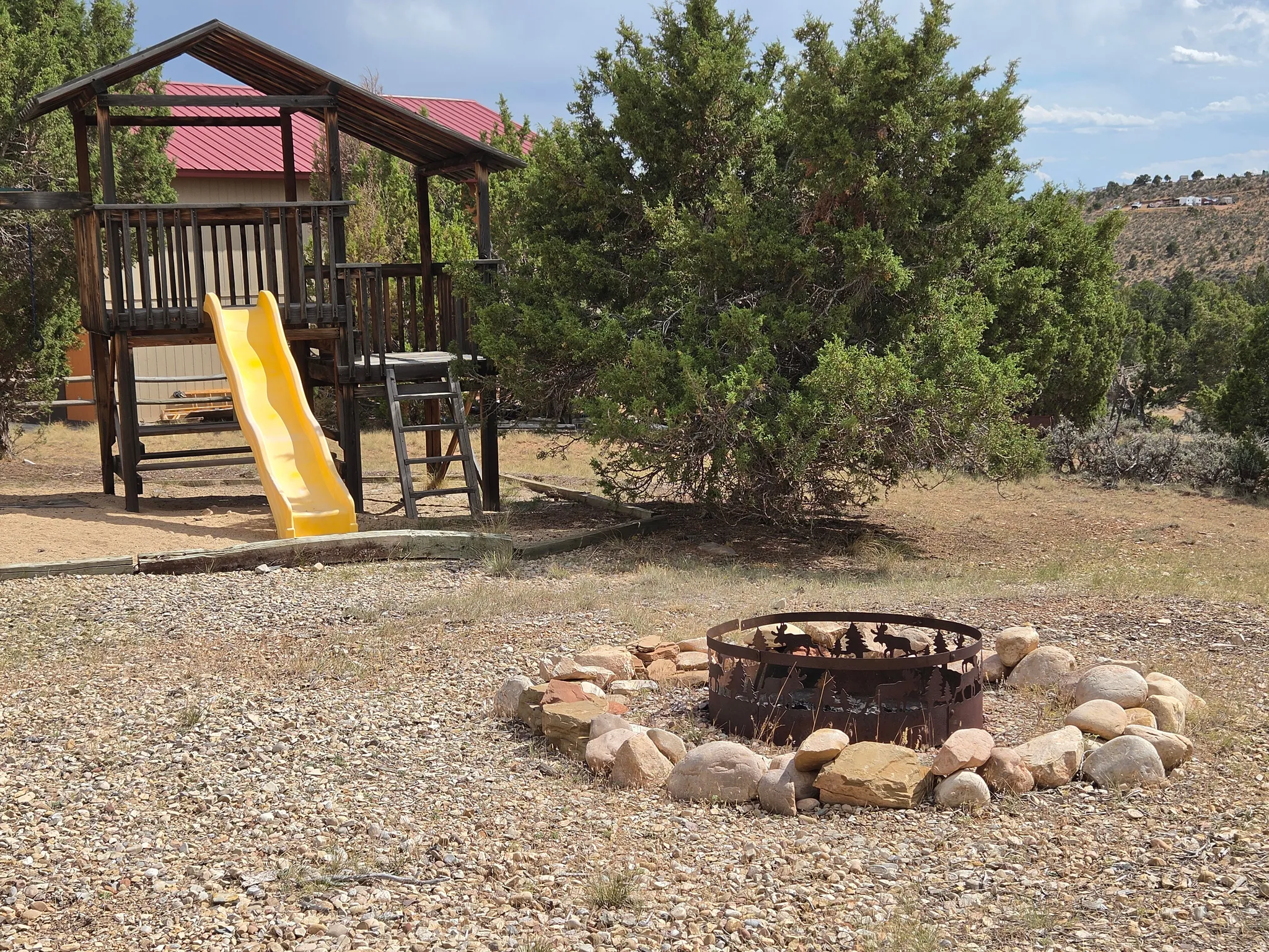 Outdoor play structure with a slide, surrounded by natural landscaping and a rock-lined fire pit