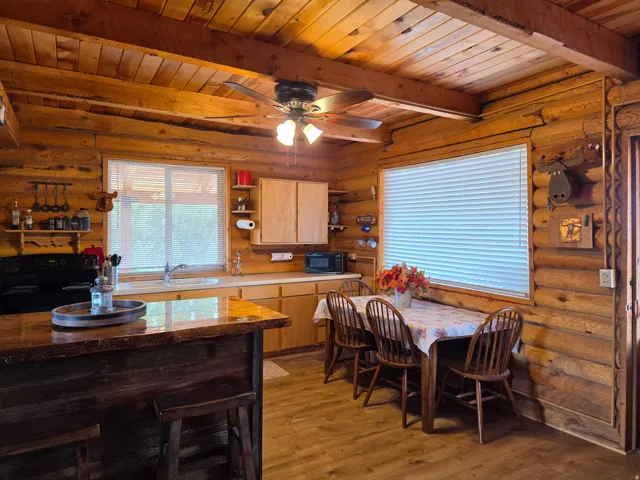 Wood-paneled walls and ceiling beams, wood-finish flooring, two large windows with blinds, a built-in kitchen island, and light wood cabinetry