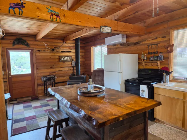 Log cabin interior featuring exposed wood beams, a prominent wood-burning stove, a wood-finish island with seating, a white refrigerator, and a black electric range