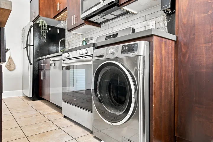 Stainless steel appliances, wood cabinetry, tile flooring, subway tile backsplash, and built-in laundry machine
