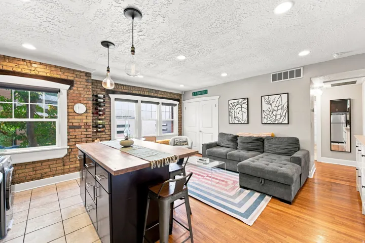 Open-concept living space featuring hardwood flooring, a dark wood-finish kitchen island with seating, brick accent wall with large windows, pendant lighting, gray upholstered sectional sofa, recessed ceiling lights, and direct access to additional rooms
