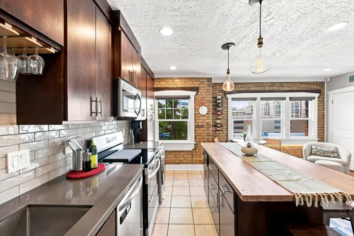 Kitchen featuring a wood-finish island, dark wood cabinetry, stainless steel appliances, a subway tile backsplash, and exposed brick accents