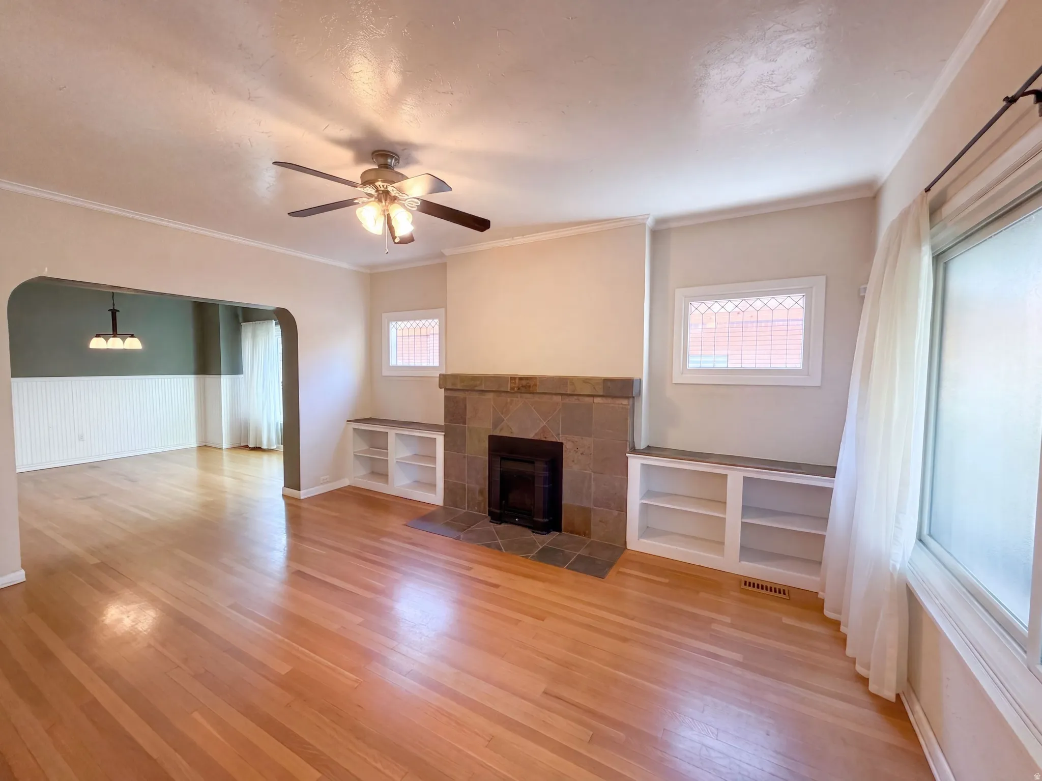 Hardwood flooring throughout, a tiled fireplace with hearth, built-in shelving, an arched doorway, and a ceiling fan with integrated lighting