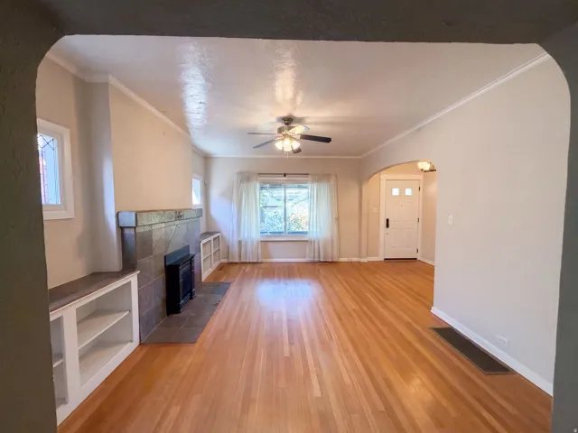 Hardwood flooring, tiled fireplace with hearth, built-in shelving, arched doorways, and a ceiling fan
