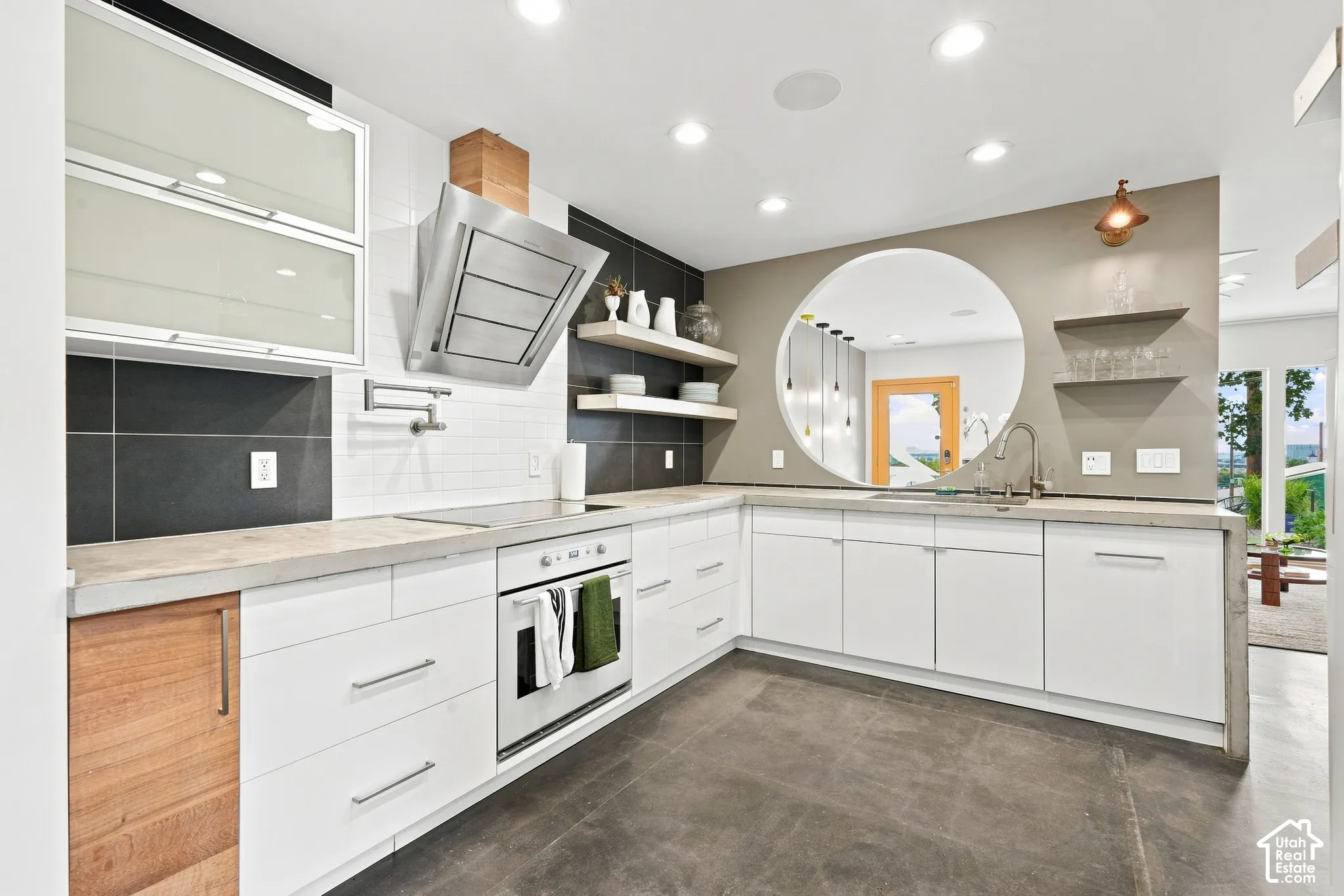 Modern kitchen featuring concrete countertops, white cabinetry, a built-in oven, stainless steel range hood, and a circular wall cut-out