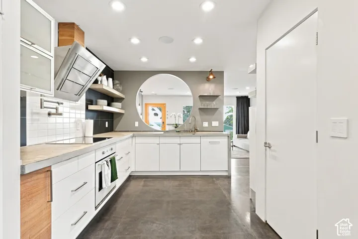 Contemporary kitchen with white cabinetry, light countertops, integrated cooktop, stainless steel range hood, and a circular wall opening