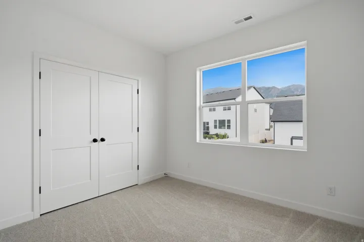 Bright secondary bedroom featuring neutral carpeting, dual white panel closet doors with black hardware, and large picture window framed with white trim allowing abundant natural light and mountain backdrop views