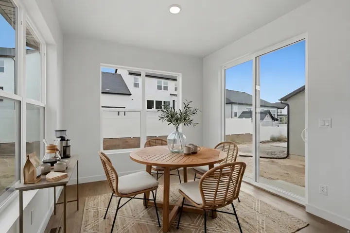 Bright dining space flooded with natural light through expansive glass doors and windows, featuring a round wood-finish table, woven accent chairs, and a neutral area rug