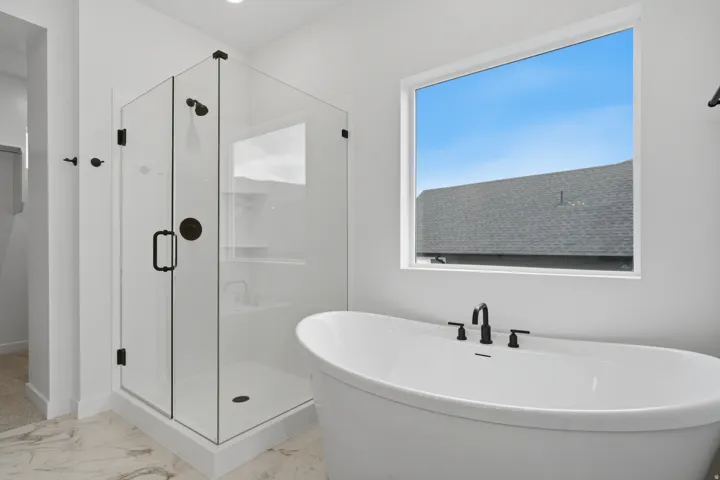 Spa-inspired primary bathroom featuring a freestanding soaking tub with matte black fixtures, frameless glass-enclosed walk-in shower with multiple showerheads, stone-finish flooring, and large picture window with natural light