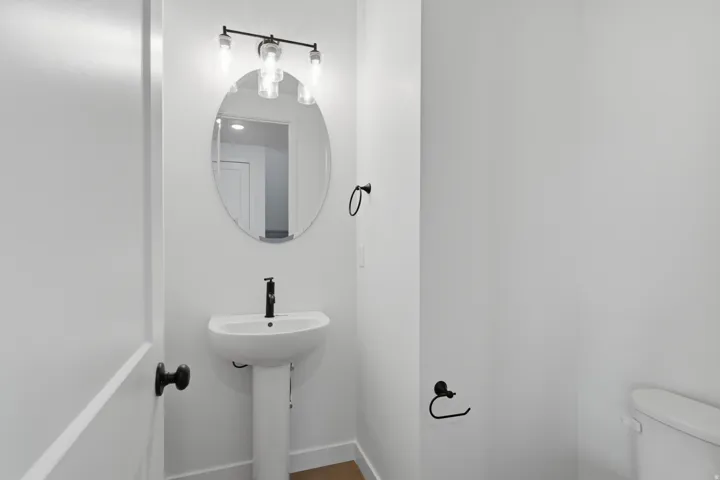 Compact powder room featuring a pedestal sink with matte black faucet, oval mirror with integrated lighting fixture, wood-finish flooring, and white painted walls with black hardware accents