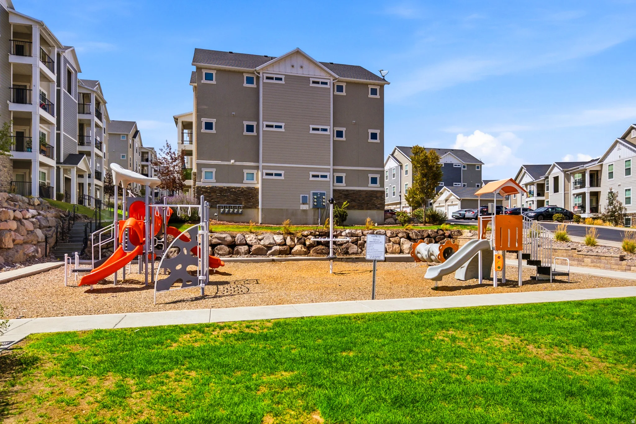 Community playground featuring dual slide structures, sandbox with natural wood edging, and manicured lawn with mature landscaping