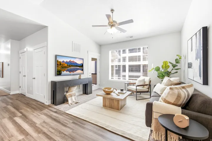 Bright, open living space showcasing wood-finish flooring, a ceiling fan with integrated lighting, and large multi-pane windows allowing natural light throughout