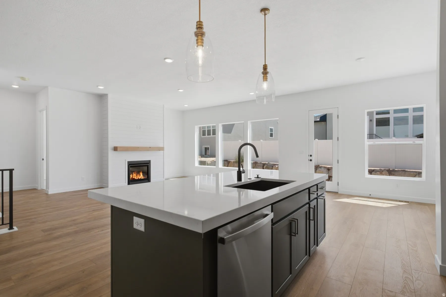Kitchen island with a light-toned countertop and integrated sink, stainless steel dishwasher, wood-finish flooring, white brick fireplace with a wood mantel, and pendant lighting fixtures
