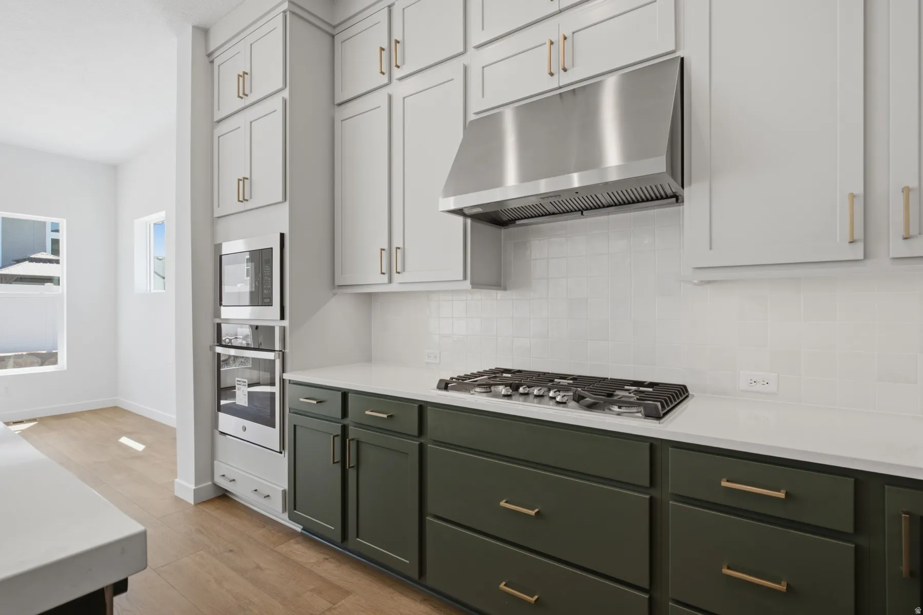 Wood-finish flooring, dual-tone cabinetry with brass hardware, stainless steel range hood, built-in double oven, and white subway tile backsplash