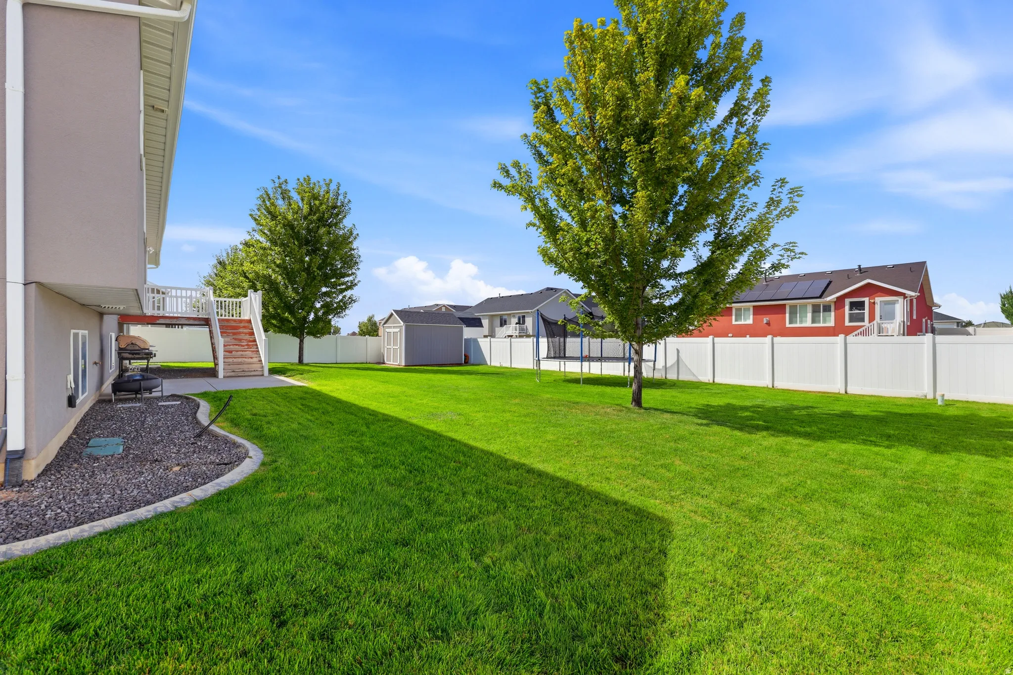Expansive backyard featuring a manicured lawn, mature trees, white privacy fencing, a wood deck with stairs, and a detached shed