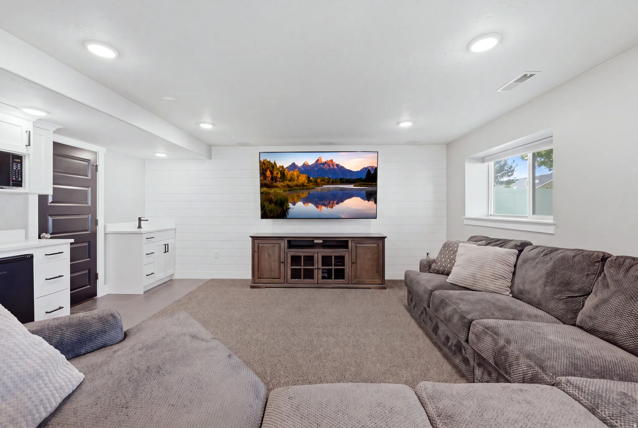 Recessed lighting, shiplap accent wall, built-in entertainment console, wet bar with white cabinetry, and a compact window