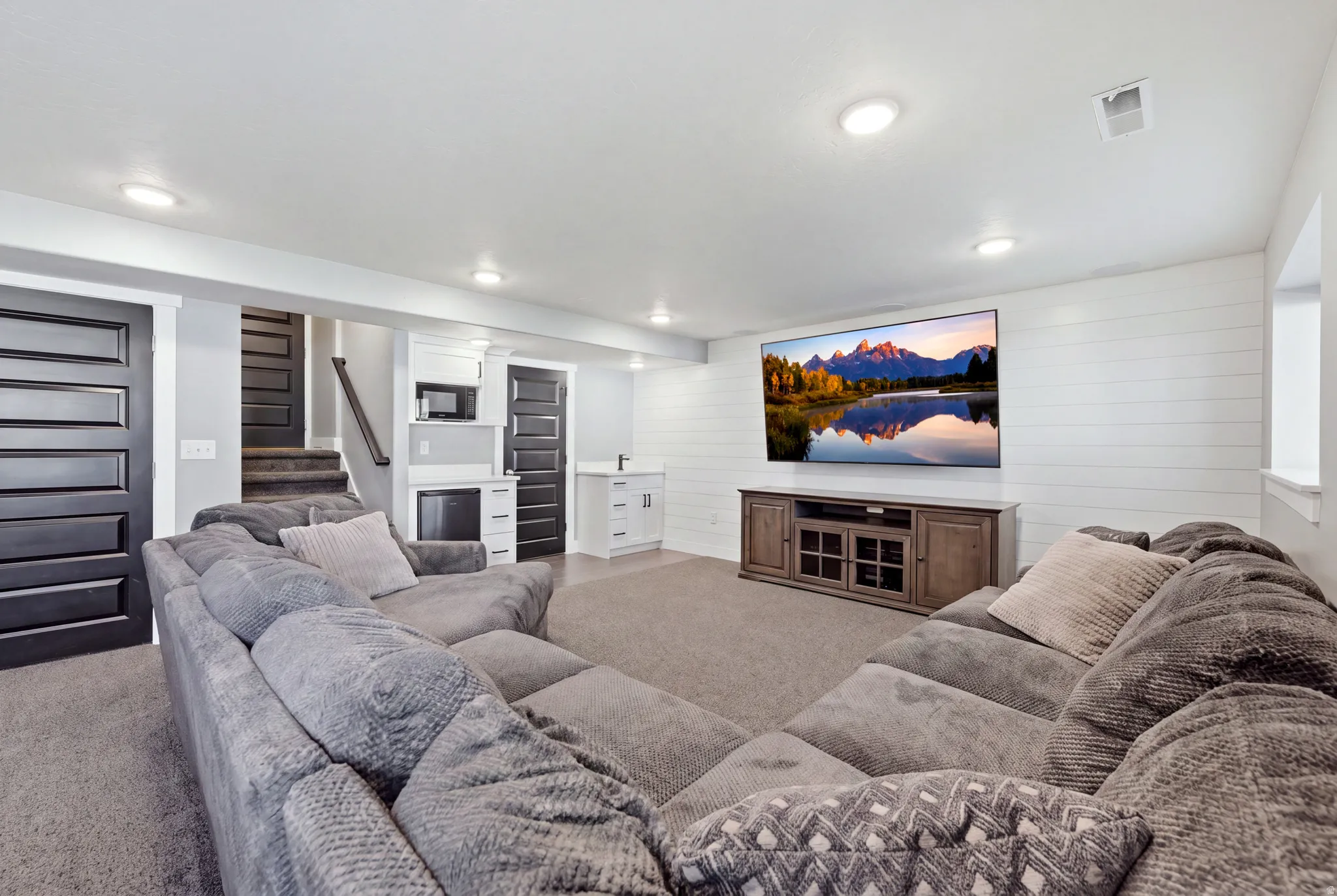Recessed lighting illuminates the carpeted living area with a built-in wet bar, white shiplap accent wall, and dark paneled doors