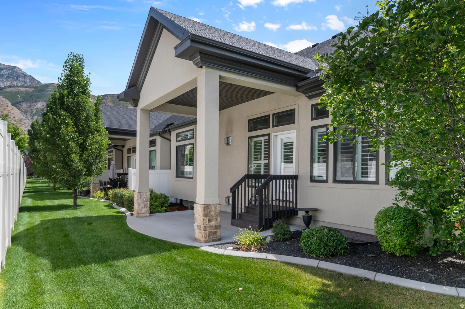 Covered patio with stacked stone pillar bases, dark wood-finish railing, and surrounding landscaping