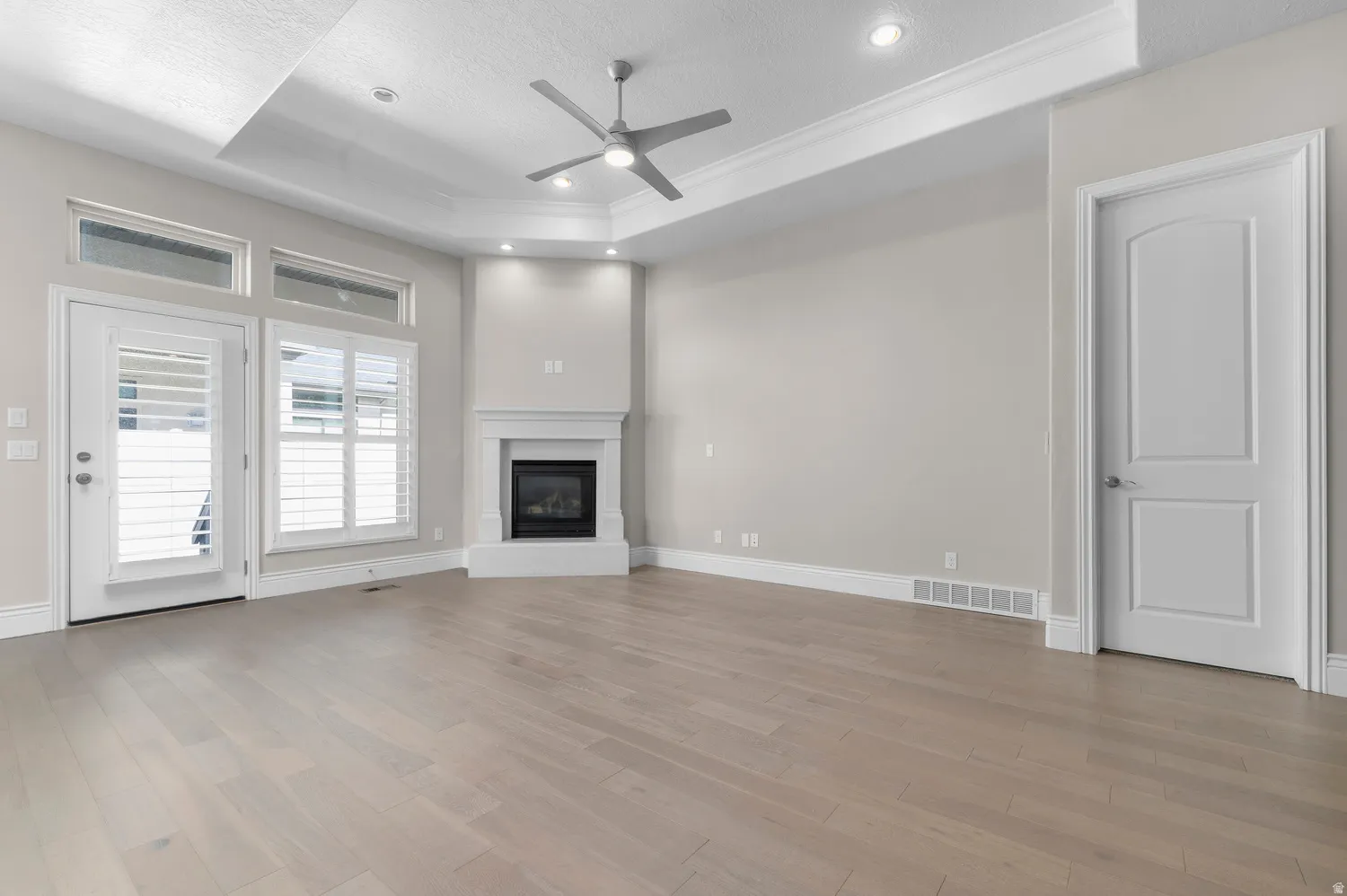 Spacious room featuring light wood-finish flooring, a tray ceiling with recessed lighting, a modern ceiling fan, a fireplace with a white mantel, and white plantation shutters on the windows