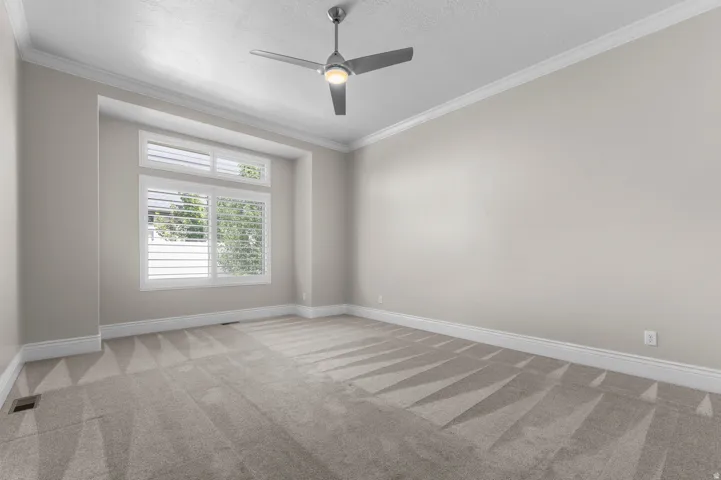 Recessed window alcove with plantation shutters, crown molding, ceiling fan with integrated lighting, and wall-to-wall carpeting