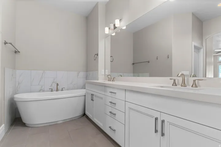 Freestanding soaking tub, white dual vanity with undermount sinks, stone countertops, large wall-mounted mirror, and tile flooring