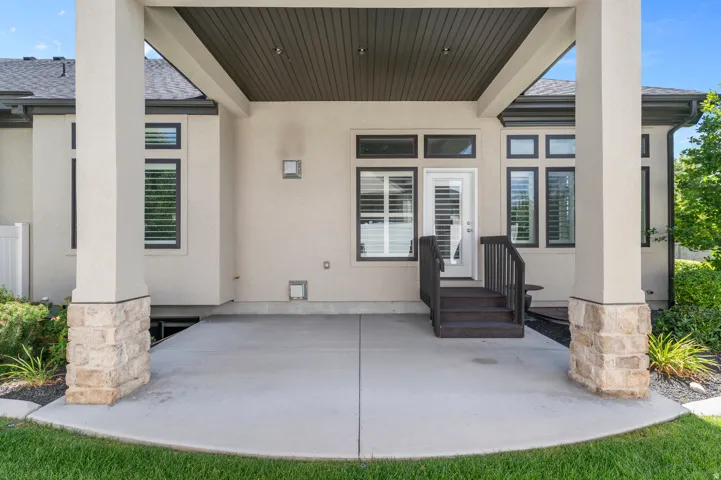 Covered patio with concrete flooring, stone-clad columns, and dark-stained steps leading to a glass-paneled door