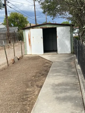 Detached metal storage structure featuring dual entry doors, concrete pathway, and chain-link fencing