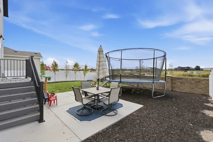 Expansive backyard featuring a concrete patio, wooden deck, manicured lawn, white privacy fence, and stone retaining wall