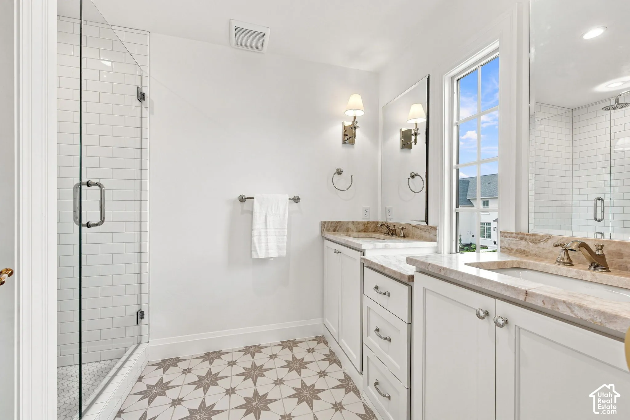 Model Home - Glass-enclosed shower with subway tile, dual vanities with integrated sinks, stone countertops, patterned floor tile, and wall-mounted sconces