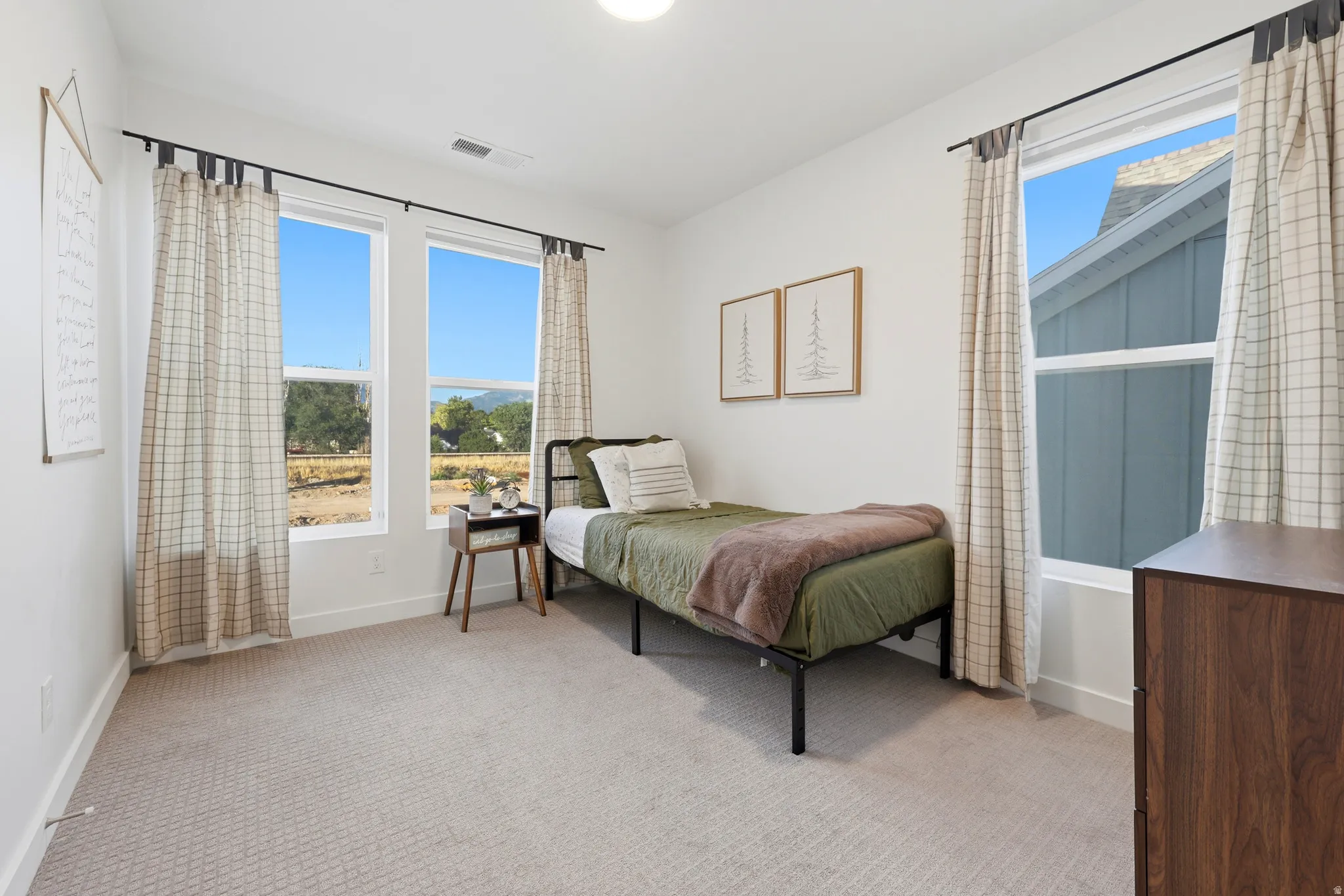 Bright secondary bedroom featuring abundant natural light from dual windows with grid-pane design, neutral carpeting, wood-finish nightstand, and access to scenic landscape vistas