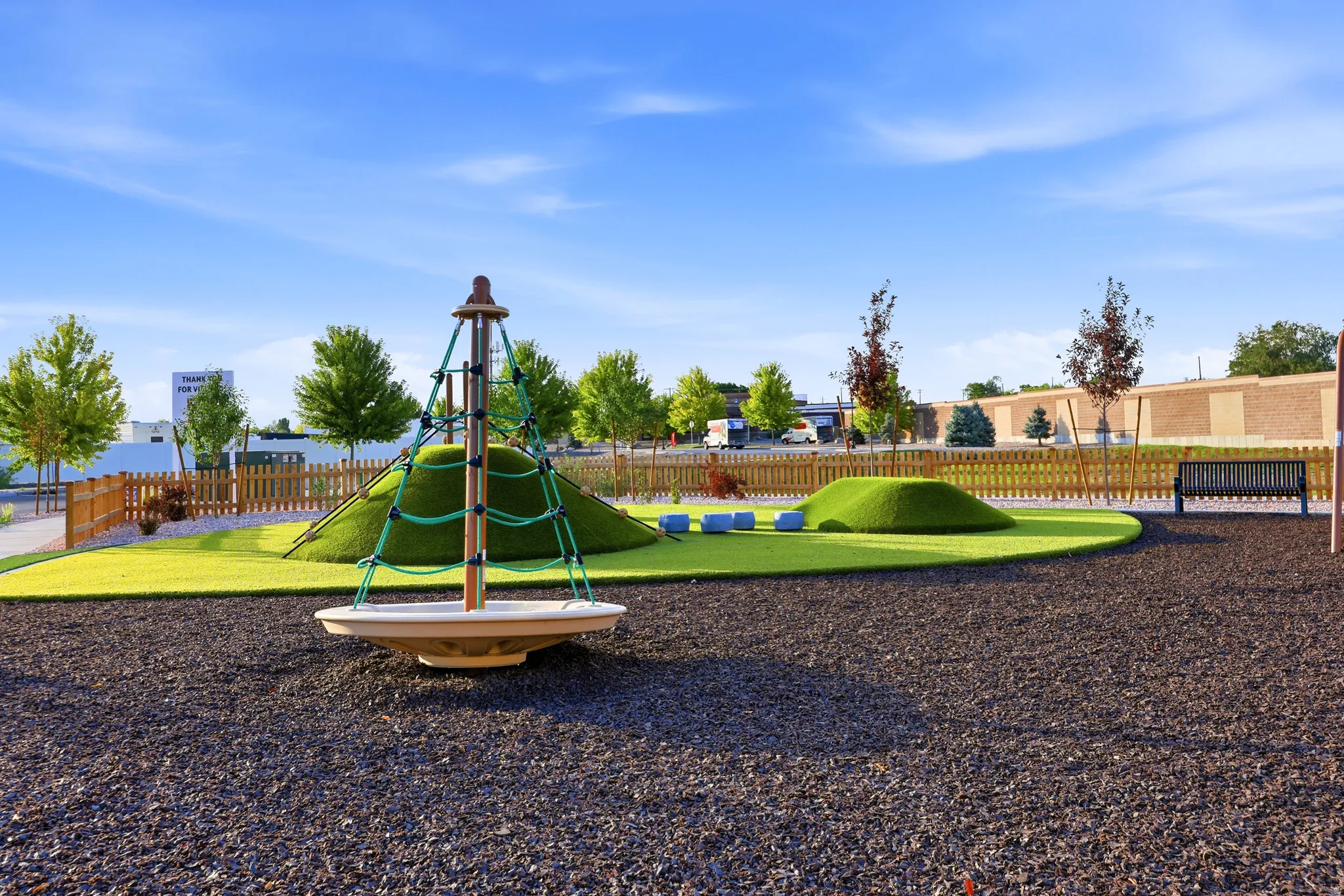 Modern recreational park featuring a multi-level playground structure with integrated slide and climbing elements, surrounded by manicured lawn areas and dark mulch surfacing