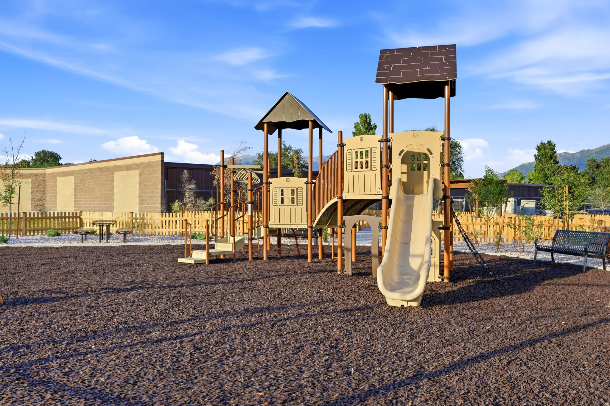 Playground structure with slides and climbing features, surrounded by woodchip ground cover, a picket fence, and a distant mountain backdrop