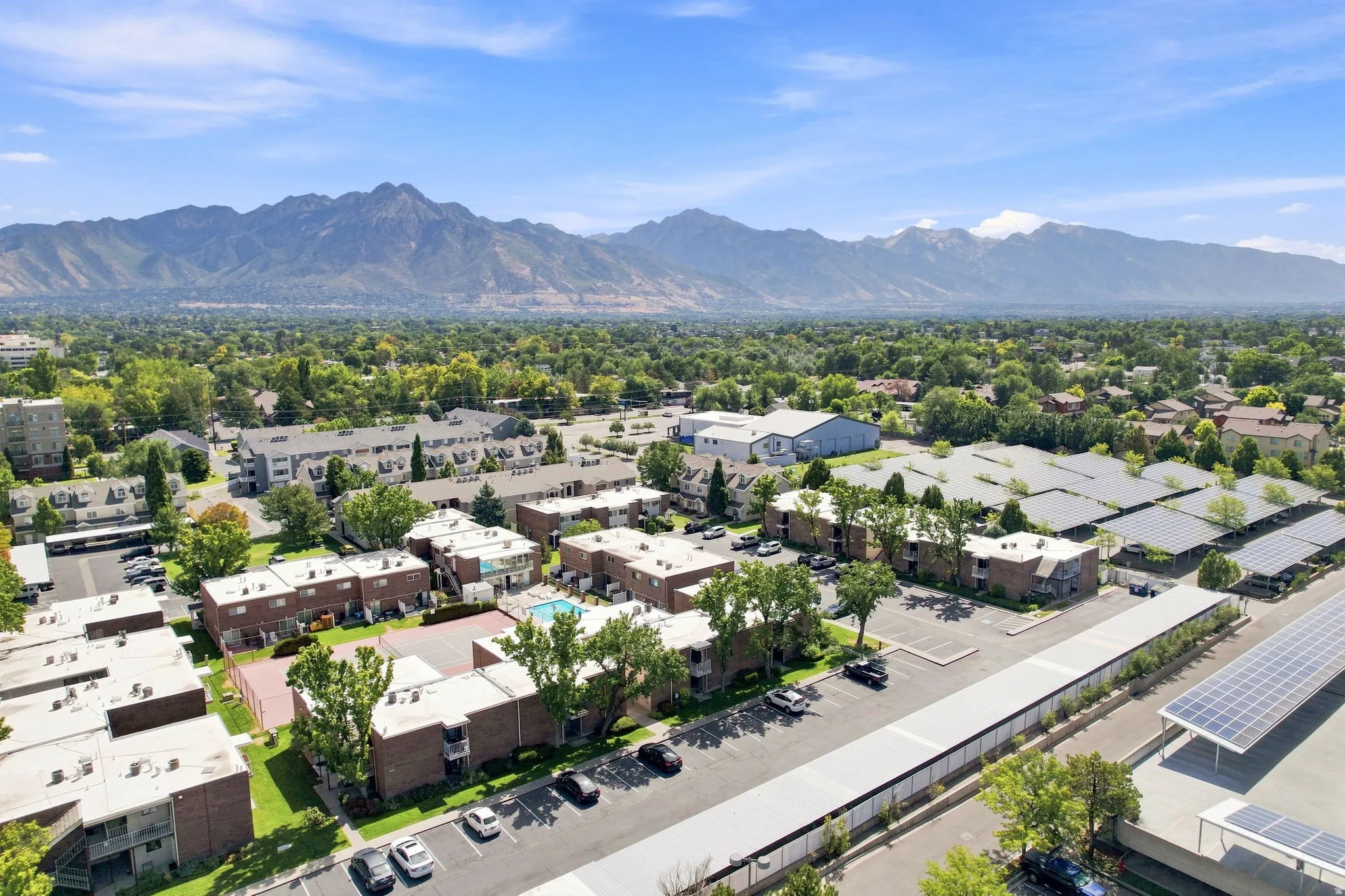Expansive residential development featuring multiple low-rise buildings, mature trees, mountain backdrop, paved parking areas, and solar panel arrays