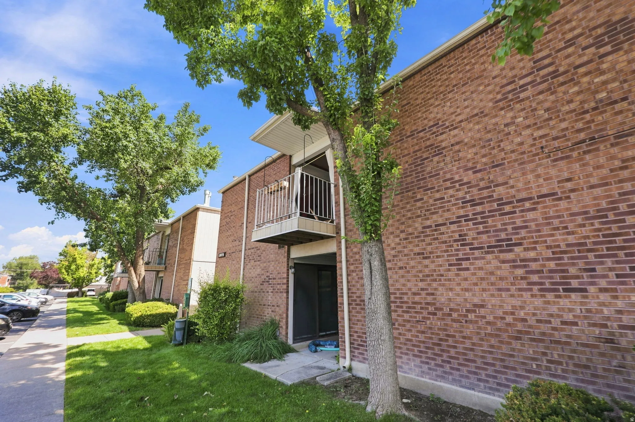 Brick exterior with a second-story balcony featuring a metal railing, mature trees, and a concrete pathway
