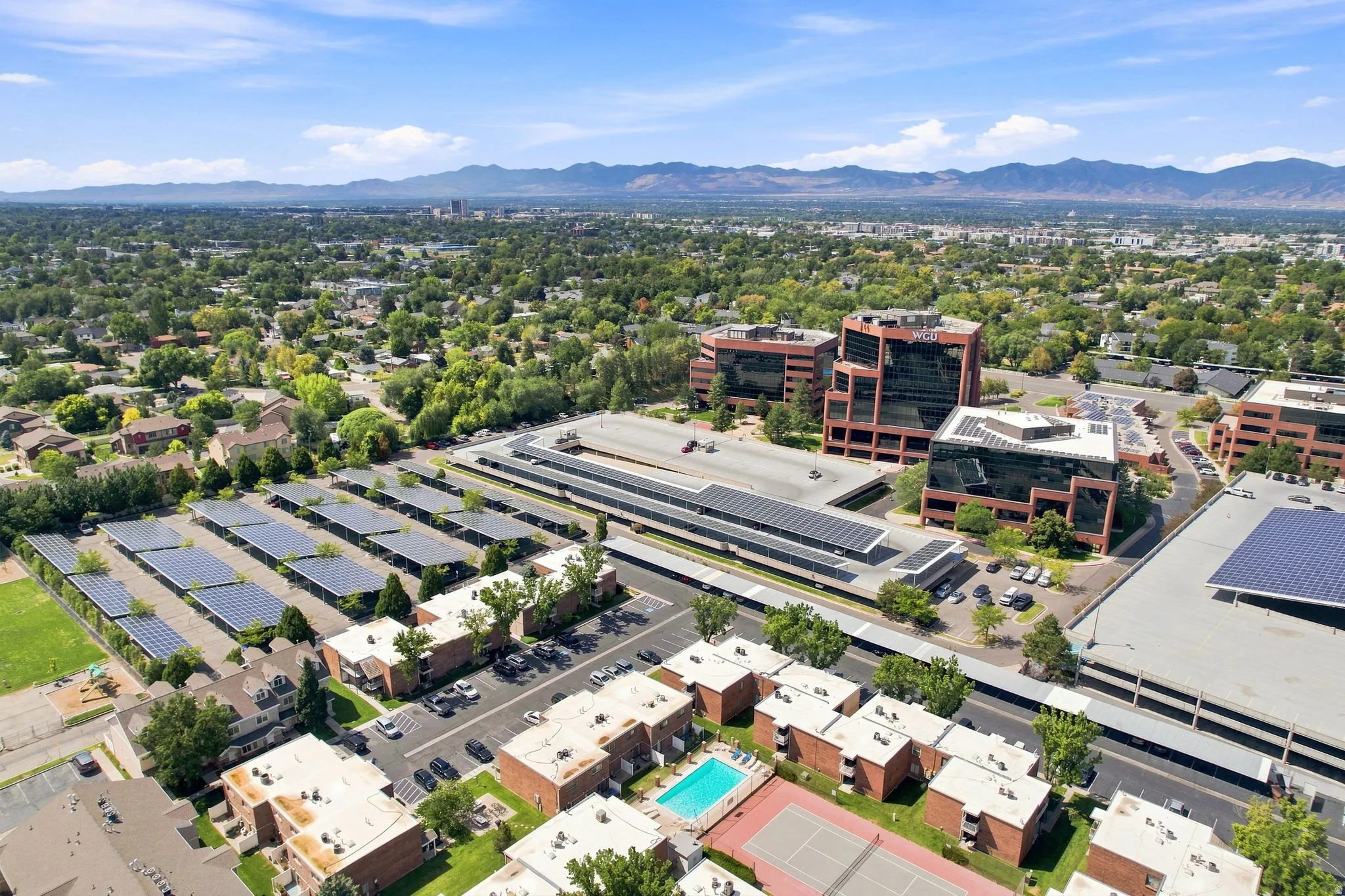 Expansive aerial perspective showcasing varied architectural styles, a large parking structure with solar panels, a residential complex with a swimming pool, and a tennis court