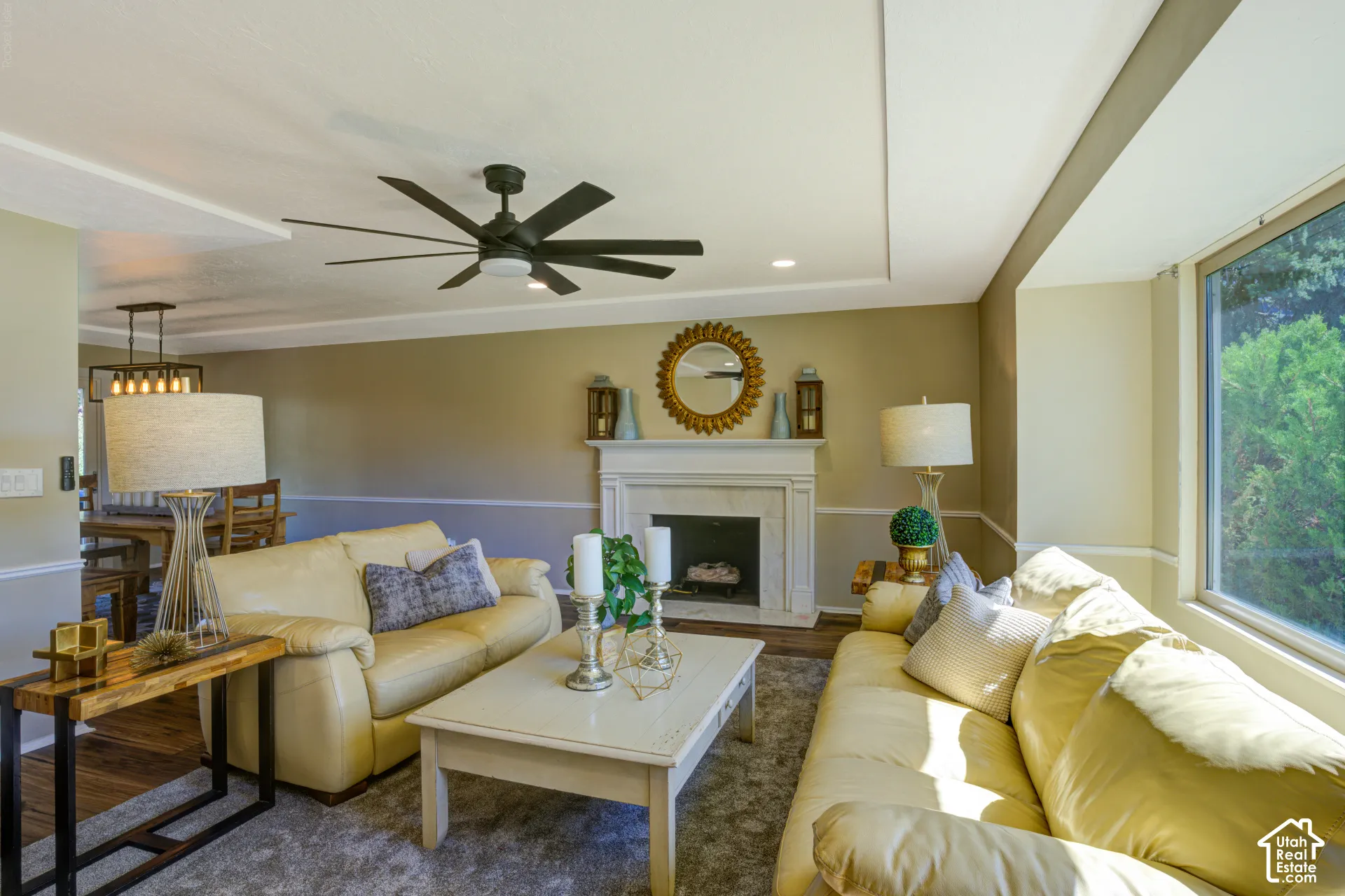 Living area featuring a fireplace, a large window, a tray ceiling, a modern ceiling fan, and hardwood flooring in the background