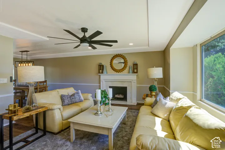 Living area featuring a fireplace, a large window, a tray ceiling, a modern ceiling fan, and hardwood flooring in the background