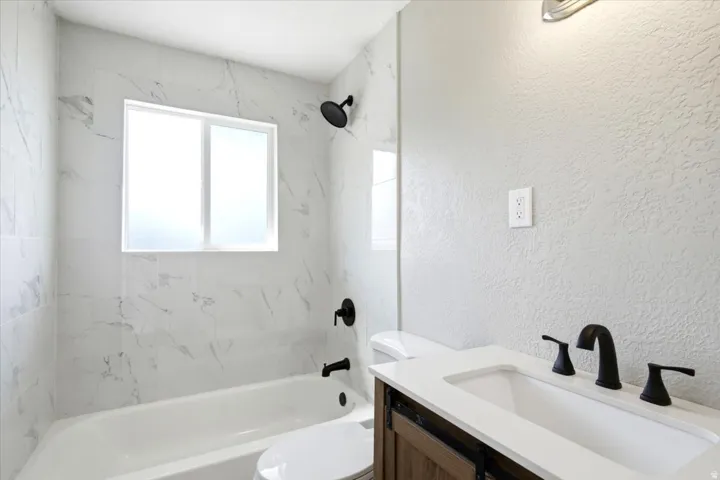 Bathtub with tile surround, frosted window, black shower fixtures, wood-finish vanity, and integrated sink
