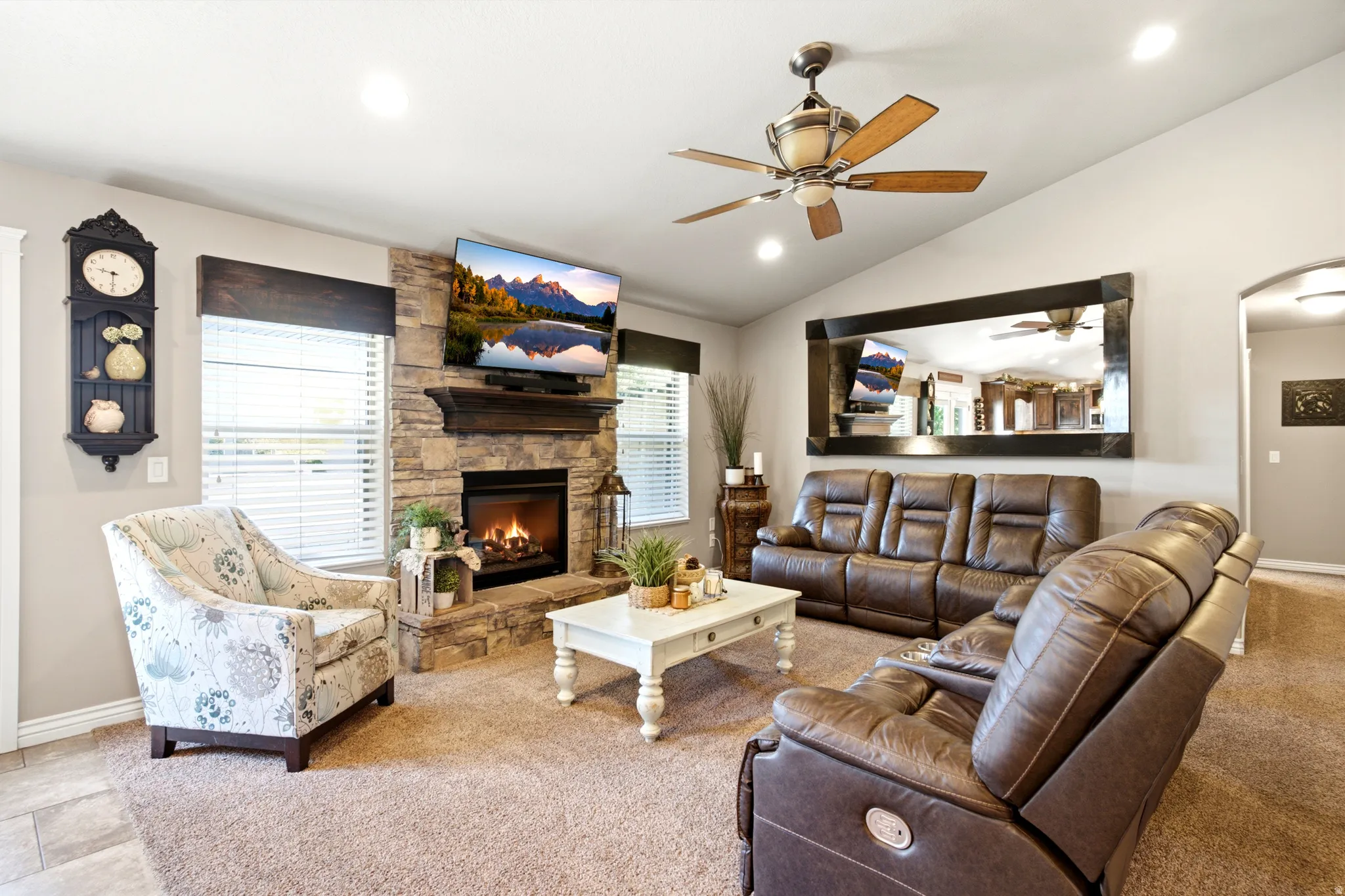 Vaulted ceiling with a ceiling fan, stone fireplace with a wood mantel, recessed lighting, and large windows with blinds