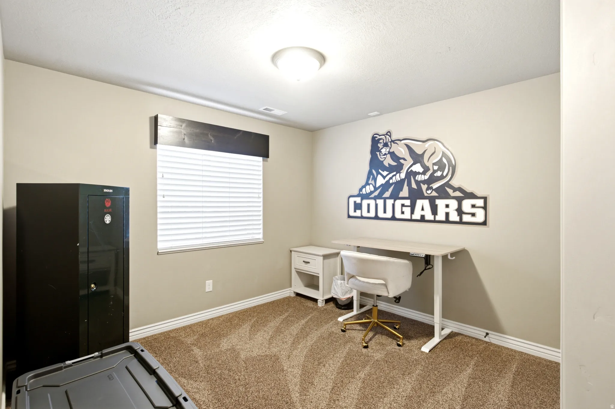 Versatile home office featuring carpet flooring, a wall-mounted desk with storage shelving, large window with horizontal blinds, recessed ceiling lighting, and branded wall decal accent