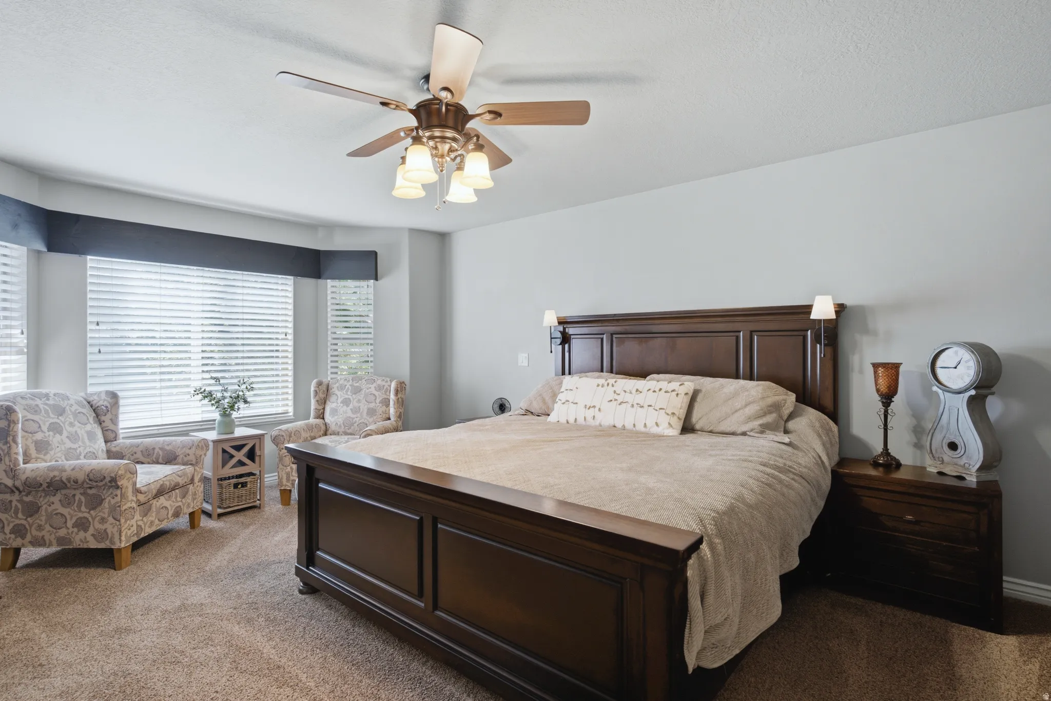 Tray ceiling with a ceiling fan, bay window with blinds, and wall-mounted sconces