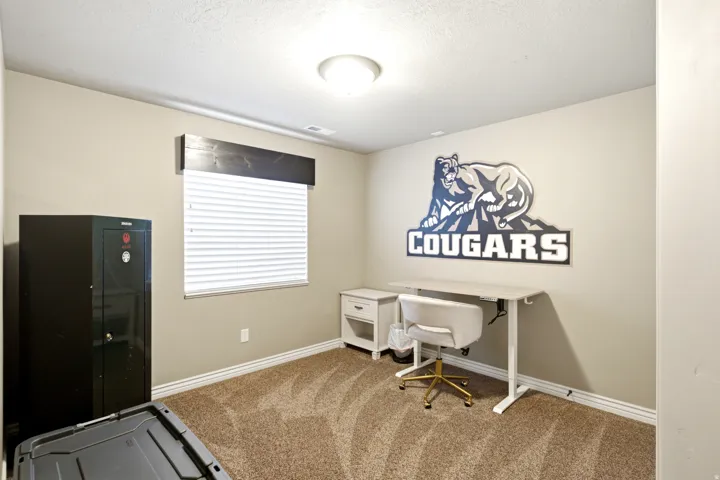 Versatile home office featuring carpet flooring, a wall-mounted desk with storage shelving, large window with horizontal blinds, recessed ceiling lighting, and branded wall decal accent
