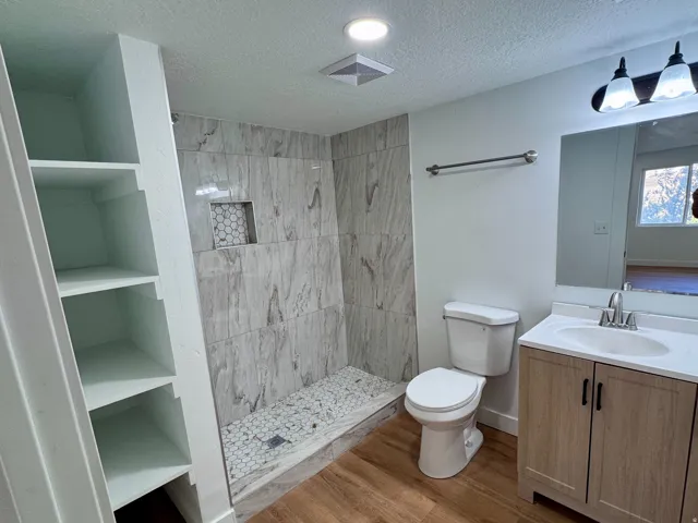 Bathroom featuring a walk-in shower with tile surround, built-in shelving, wood-finish flooring, and a single vanity with a white countertop