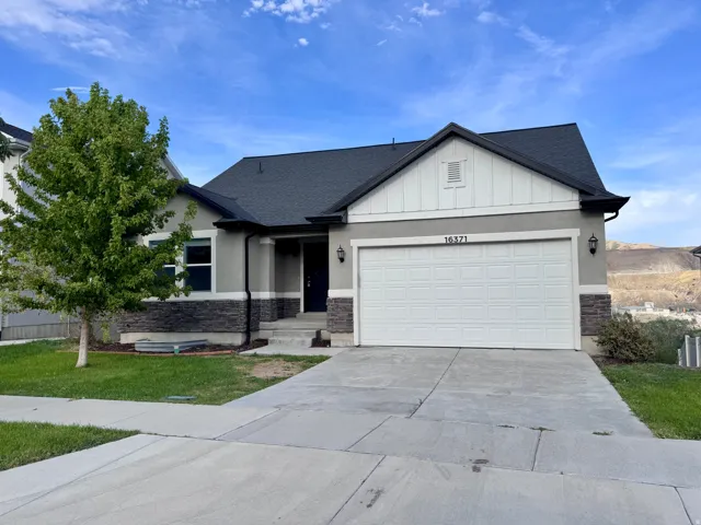 Stucco exterior with stone accent, attached two-car garage, dark asphalt shingle roof, paved driveway, and established front landscaping