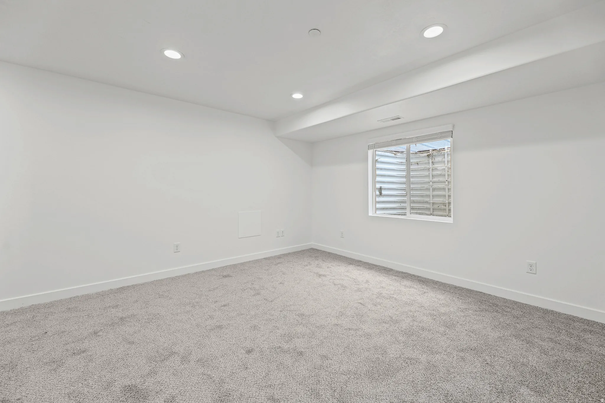 Recessed ceiling lighting, light gray wall-to-wall carpeting, white painted walls, and a window with a window well