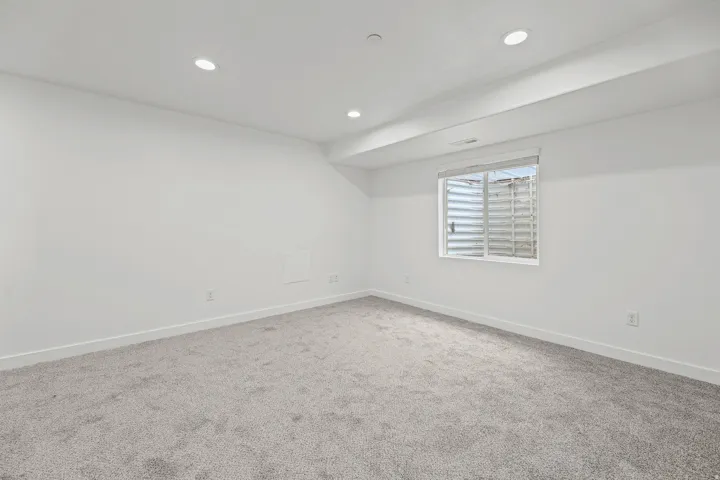 Recessed ceiling lighting, light gray wall-to-wall carpeting, white painted walls, and a window with a window well