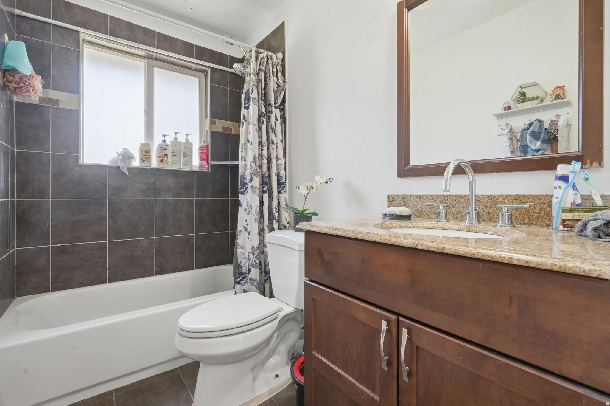 Bathtub with dark tile surround, window, white toilet, and wood-finish vanity with a light-toned countertop and integrated sink
