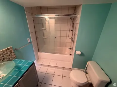 Bathroom featuring a glass vessel sink, tiled countertop, under-sink cabinetry, white tile flooring, and a shower-over-tub with glass enclosure