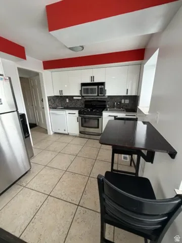 Kitchen featuring gloss white cabinetry, black stone-finish backsplash, stainless steel appliances, and tile flooring